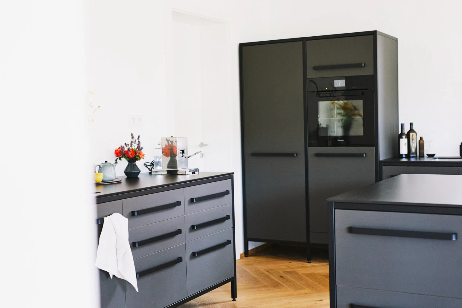 Moderne Küche in Grau und Schwarz mit Holzboden, schwarzem Einbauofen und Hochschrank.